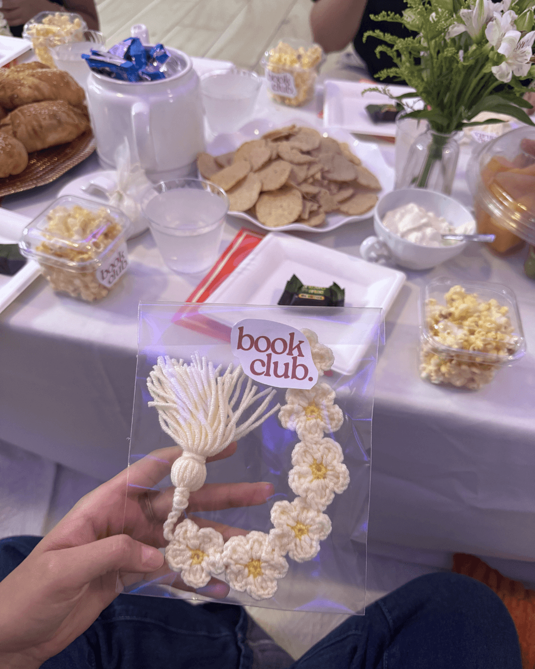 Mano sosteniendo un dije tejido a crochet con sticker de book club, snacks de fondo