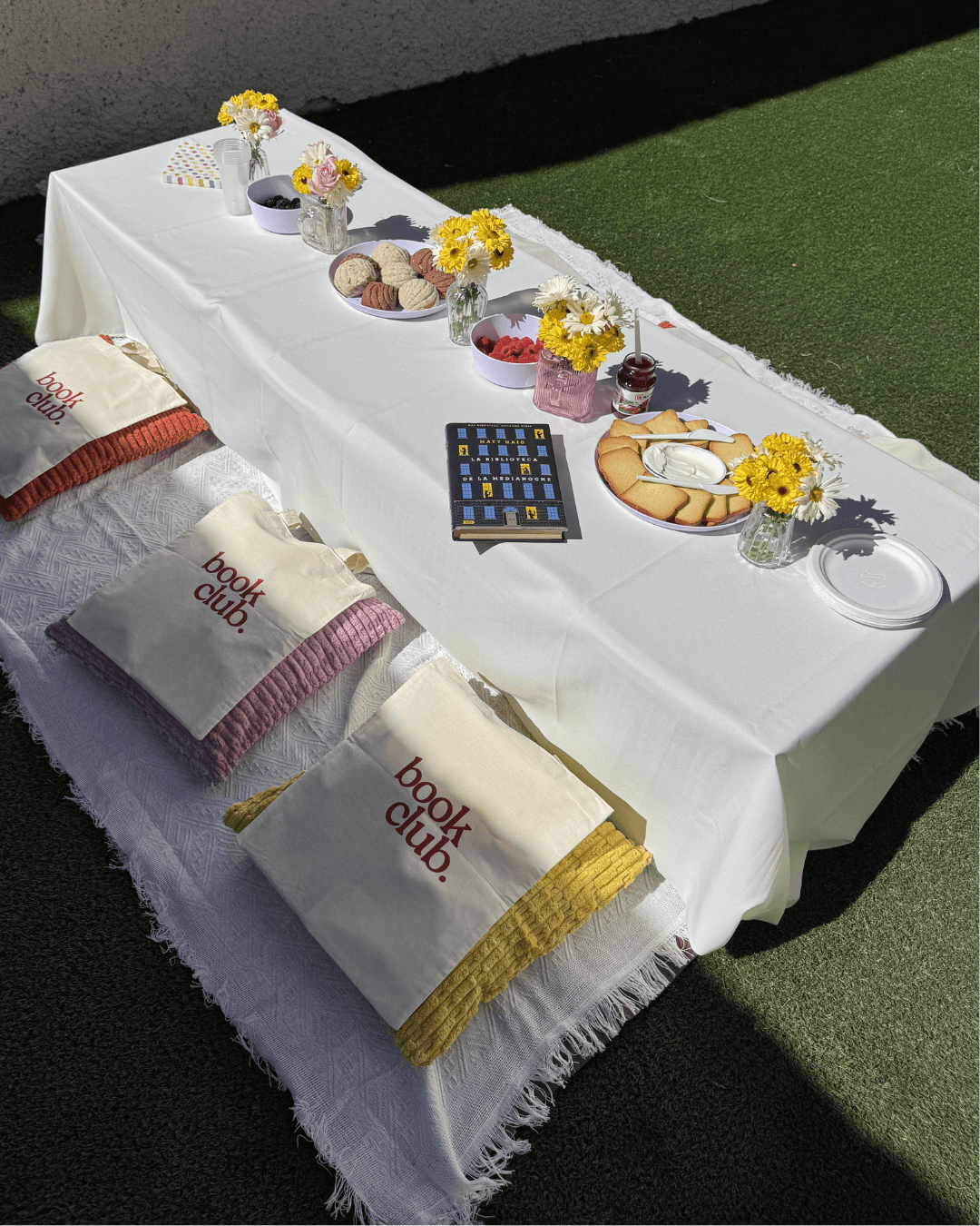Mesa de picnic con mantel blanco, tote bags de book club sobre cobijas de colores, flores y snacks